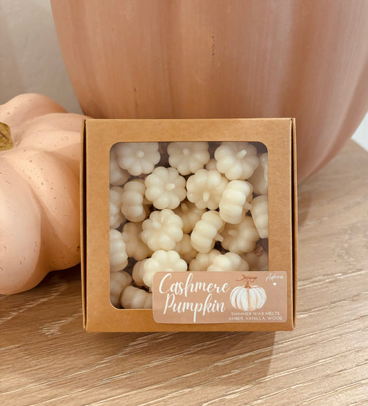A close-up of white, 3D pumpkin-shaped wax melts arranged on a rustic wooden tray. The smooth surface and delicate details of the pumpkins are highlighted, with a soft autumn background.