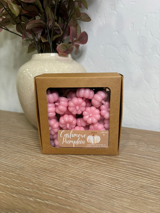 A close-up of soft pink, 3D pumpkin-shaped wax melts arranged on a rustic wooden tray. The smooth surface and delicate details of the pumpkins stand out against a warm, cozy autumn background.