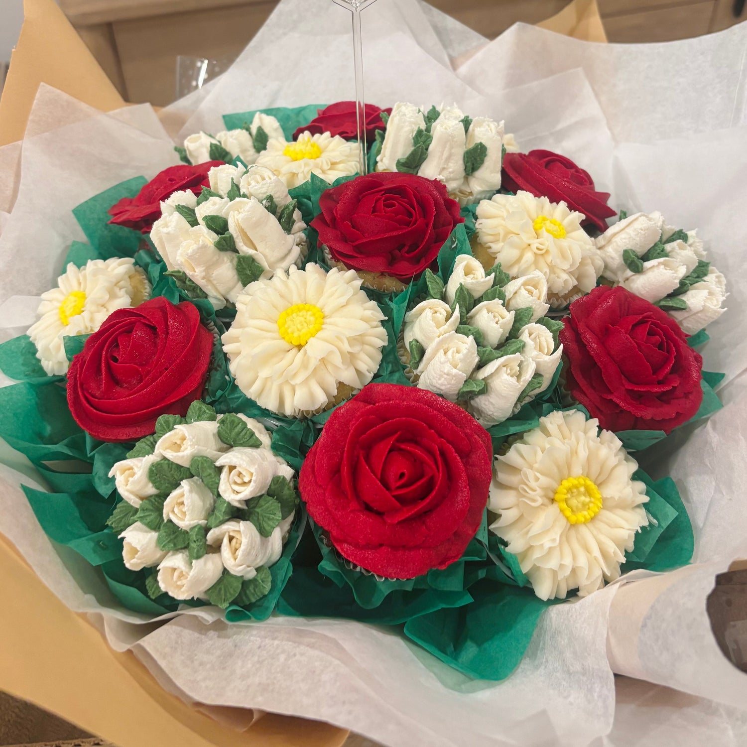 A floral bouquet made from decorated cupcakes, featuring red and white frosting roses and daisies with green frosting leaves, wrapped in white tissue paper and a clear stand with a 'thank you' card.