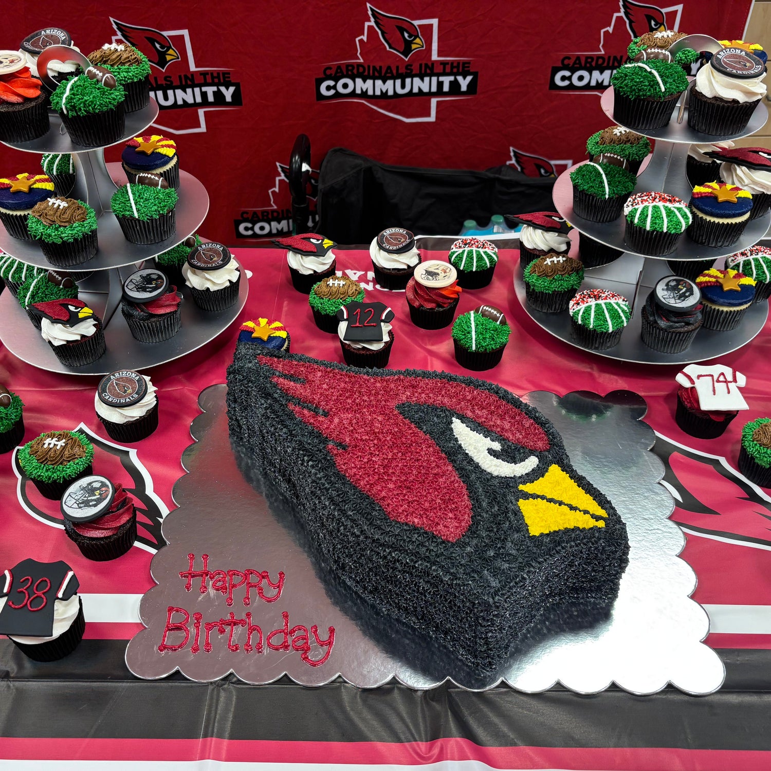 A table covered with cupcakes and cake toppers themed around the Arizona Cardinals, with a large cardinal head cake in the foreground and a red banner in the background.