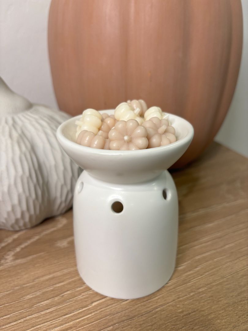 A tan and white wax melt shaped like a pumpkin beginning to melt in a wax warmer. A subtle shimmer is emerging from the melt, creating a sparkling effect as the amber-toned liquid pools.