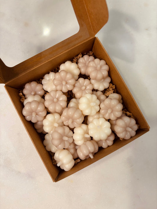 A close-up of tan and white, 3D pumpkin-shaped wax melts arranged on a rustic wooden tray. The smooth surface and intricate details of the pumpkins are highlighted, set against a warm autumn backdrop.